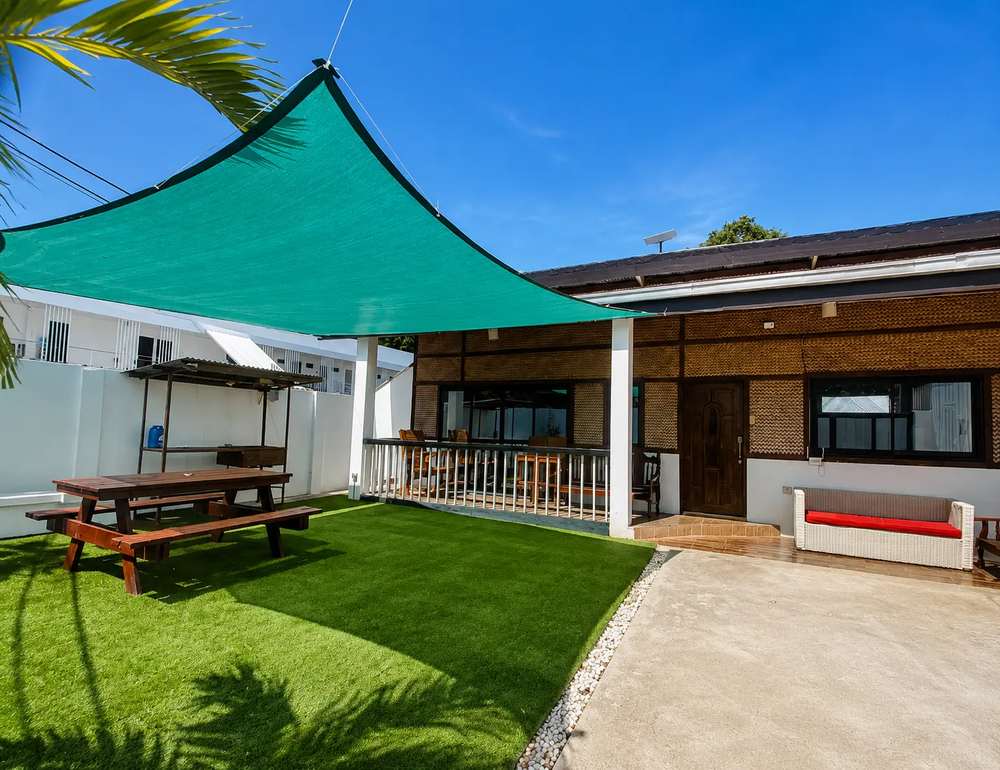 Garden courtyard at Mayumi Lodge with artificial lawn, a wooden picnic table under a green shade sail and a covered terrace with rattan seating