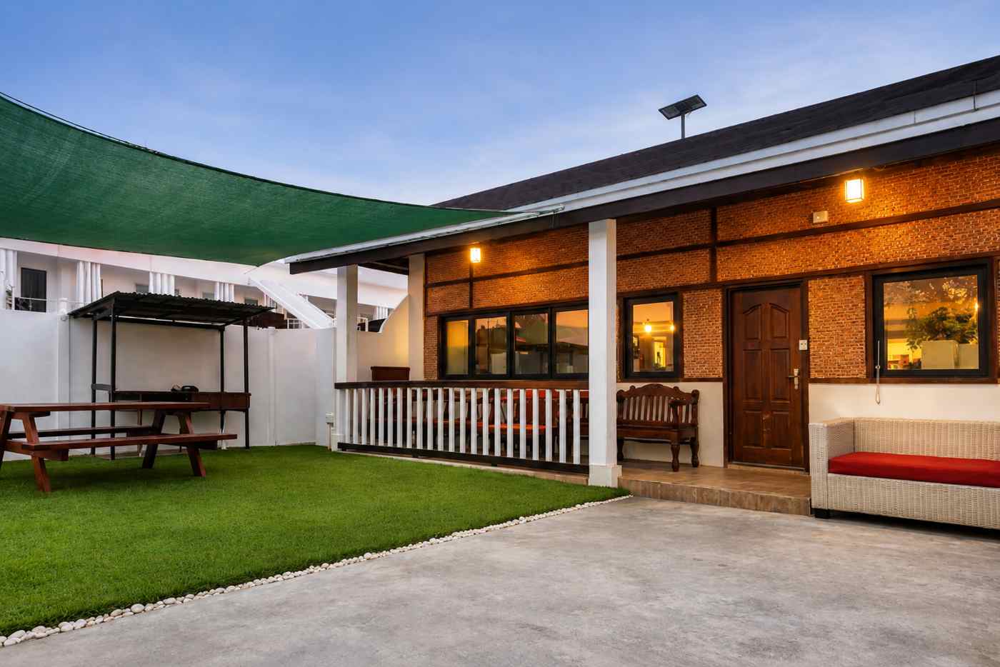 Brick-fronted family house entrance at Mayumi Lodge at dusk, with a covered porch, rattan sofa and lawn seating in front